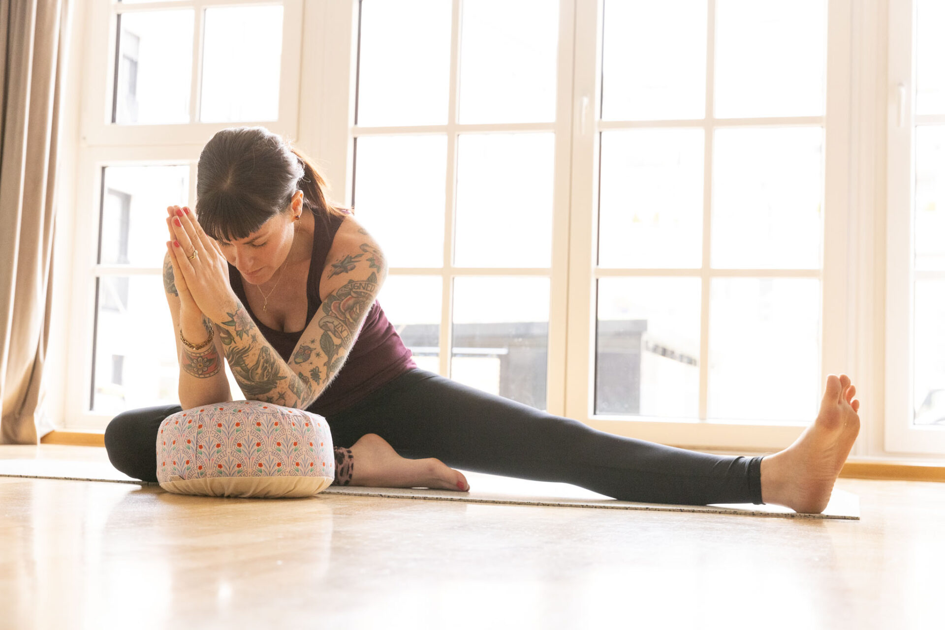 Laura Köppen vom Yogastudio Place to Be in München sitzt in einer tiefen Yin Haltung auf der Matte. Ihre Arme hat sie auf ein Meditationskissen aufgestützt, der Kopf ist nach unten gesenkt, die Augen sind geschlossen. Ein Bein ist abgewinkelt, das andere ausgestreckt.